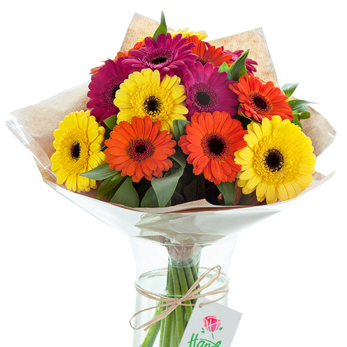 In this richly detailed image, a festive hand-tied bouquet of Gerbera daisies glows with Battersea's signature vivacity. The bouquet, gracefully poised in a clear, slightly hourglass-shaped vase, features a dynamic mix of large blooms-sunny yellow, bold orange, and striking magenta-each showcasing velvety petals and a distinctive black-brown center. Sprays of lush green foliage wind through the blossoms, adding freshness and volume reminiscent of flower stalls near Clapham Junction or along St. John's Hill. The upper arrangement sits wrapped in textured, light brown kraft paper, softly folded to cradle the flowers, with a crinkled layer of clear cellophane arching outward in a protective embrace. Through the pristine transparency of the vase, sturdy green stems are visible, tidily gathered by a piece of natural raffia or twine. At the centre of this bundle, a small rectangular white gift tag swing from the knot-its red rose logo and partial handwritten note adding a hint of bespoke artistry, as if left by a thoughtful florist prepping the bouquet for a special moment in Battersea. The whole presentation, with its interplay of radiant colors, textural depth, and gentle lighting, evokes happiness and appreciation-an ideal choice for celebrating milestones, sending good wishes, or brightening a loved one's day nearby. This floral arrangement tells a story of skilled hands, community spirit, and the everyday beauty that local Battersea florists bring to life.